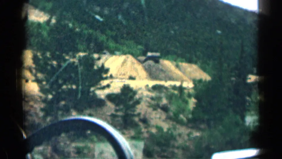 Interior passenger-view 8mm frame in Colorado with windshield edge, steering wheel, rocky roadside, and a car ahead.