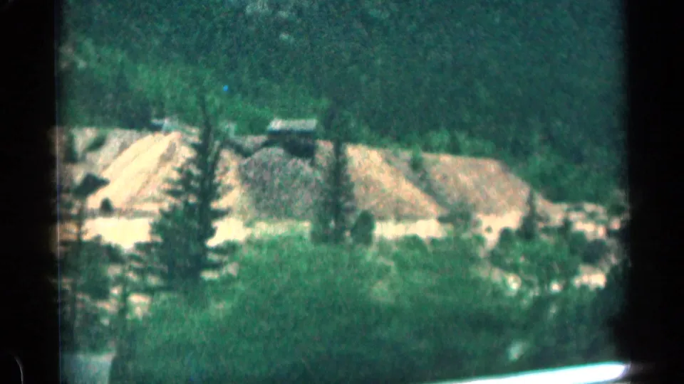 Colorado mountain roadside with pale gravel or sand piles, trees, and steep green slopes, filmed from a passing car in 1963.