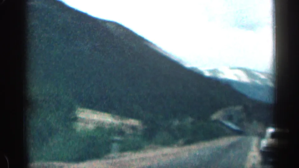 Passenger-view frame of a winding Colorado mountain road with forested slopes and distant pale sky, from 1963 8mm home-movie footage.