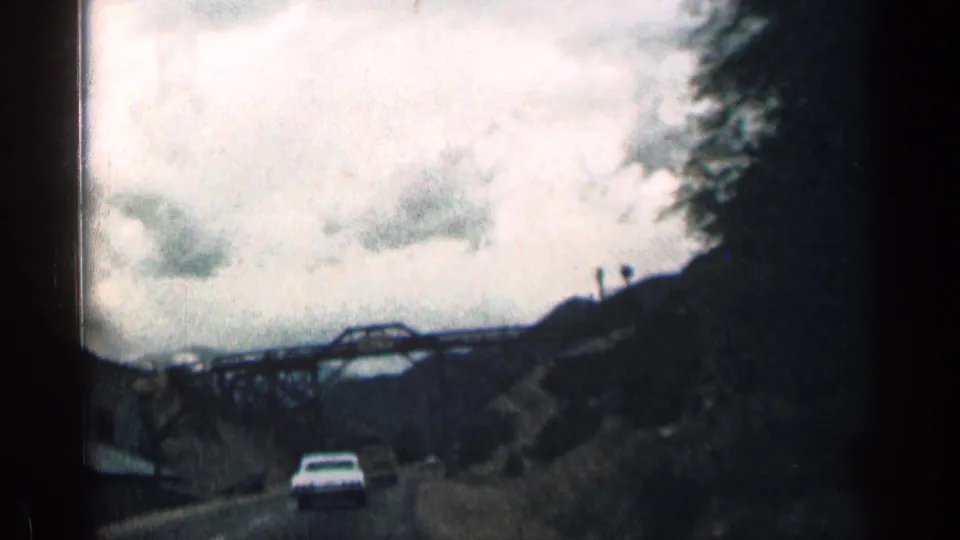 A white car drives toward a metal truss bridge on a Colorado mountain road, with dark hillsides and cloudy sky in 1963 8mm footage.