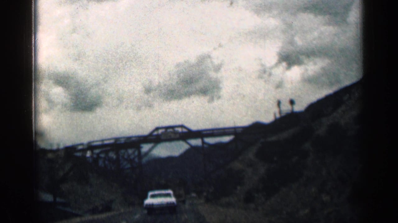 White car approaches a steel truss bridge beneath a cloudy sky.