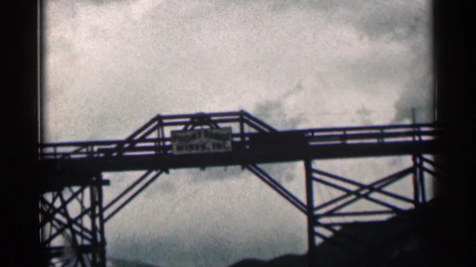 Closer 1963 Colorado frame of a steel bridge sign appearing to read Front Range Mines, Inc. against a cloudy sky.