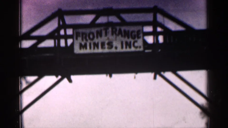 Close 8mm frame of a steel bridge sign appearing to read Front Range Mines, Inc., with truss beams silhouetted around it.