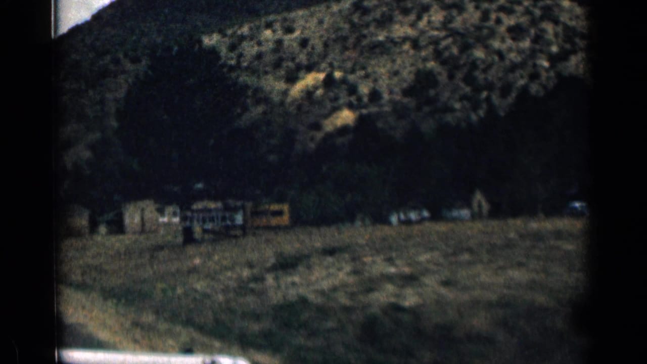 1963 Colorado 8mm frame showing a shadowed mountain roadside seen through a moving car, with distant structures and parked vehicles.