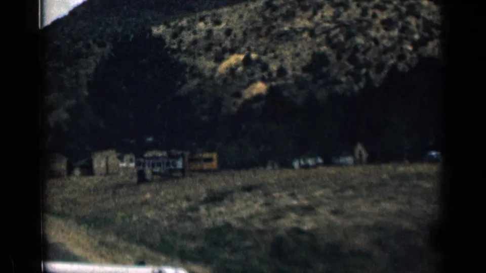 1963 Colorado 8mm frame showing a shadowed mountain roadside seen through a moving car, with distant structures and parked vehicles.