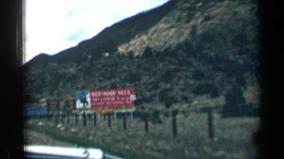 1963 Colorado highway view showing a large Berthoud Pass billboard, fence posts, and rocky mountain slope from a moving car.