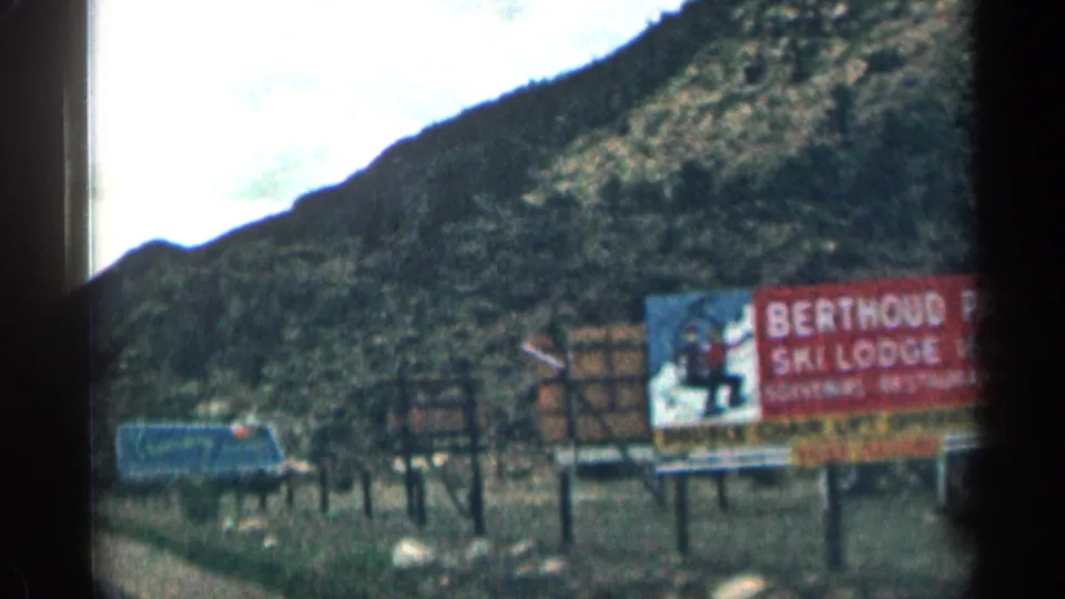 1963 Colorado frame showing a Berthoud ski lodge roadside billboard, nearby vehicles, fence posts, and rugged mountain terrain.