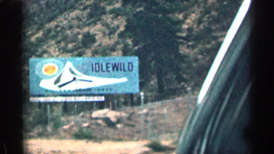 1963 Colorado 8mm frame of an Idlewild billboard beside a rocky roadside, seen past a car window frame.