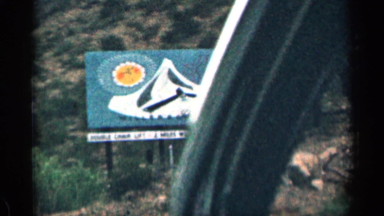 1963 Colorado roadside billboard with a stylized mountain and sun, partially obscured by the dark frame of a moving car window.