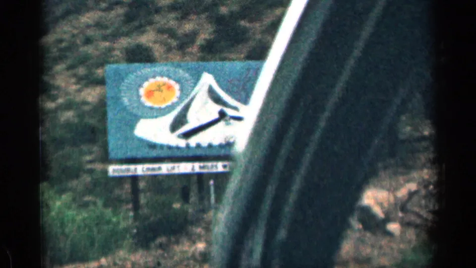 1963 Colorado roadside billboard with a stylized mountain and sun, partially obscured by the dark frame of a moving car window.