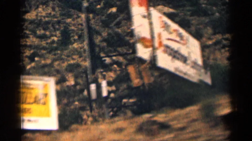 Motion-blurred 1963 Colorado roadside signage, including a tilted billboard face, yellow sign edge, posts, and dark hillside terrain.