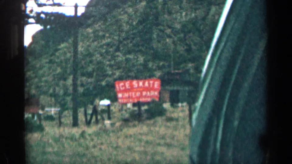 1963 Colorado 8mm frame showing a red ice skate rental sign near trees and hills beside a road.