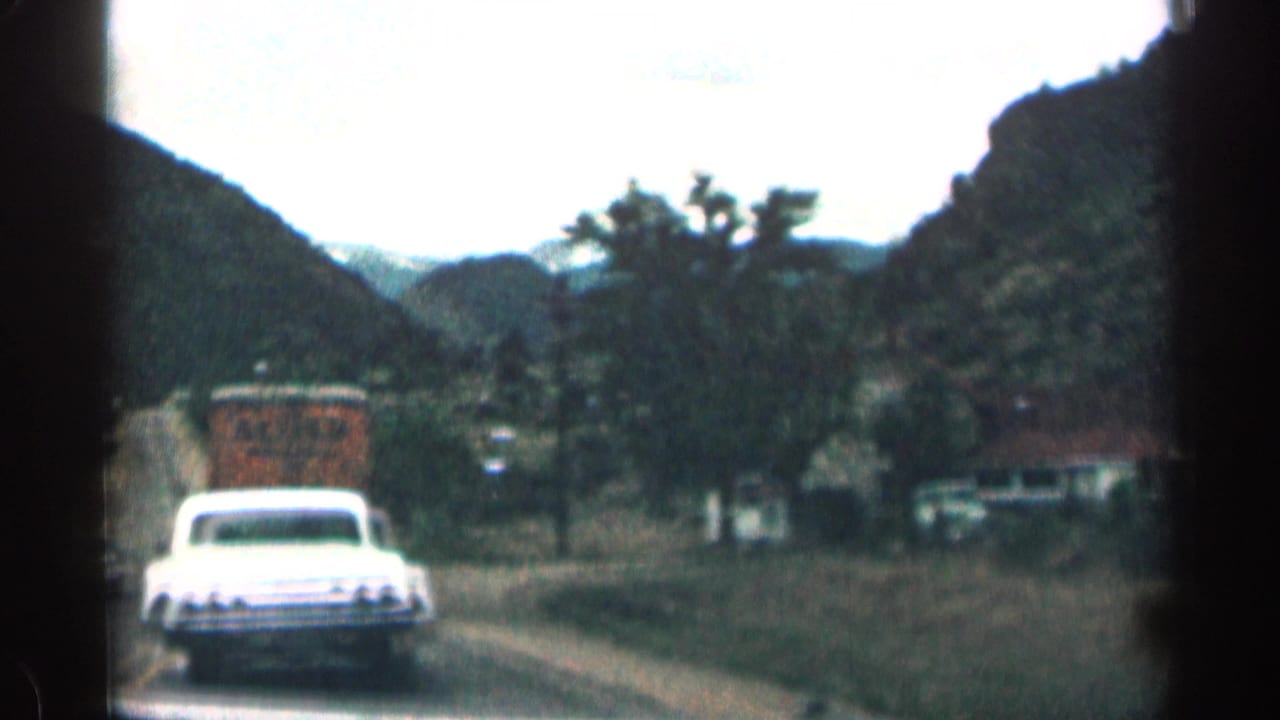 White car follows a truck on a narrow road through mountains.