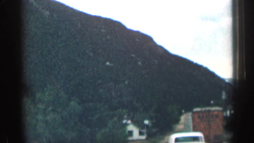 1963 Colorado home-movie frame with a dark mountain slope, roadside buildings, and a vehicle ahead.