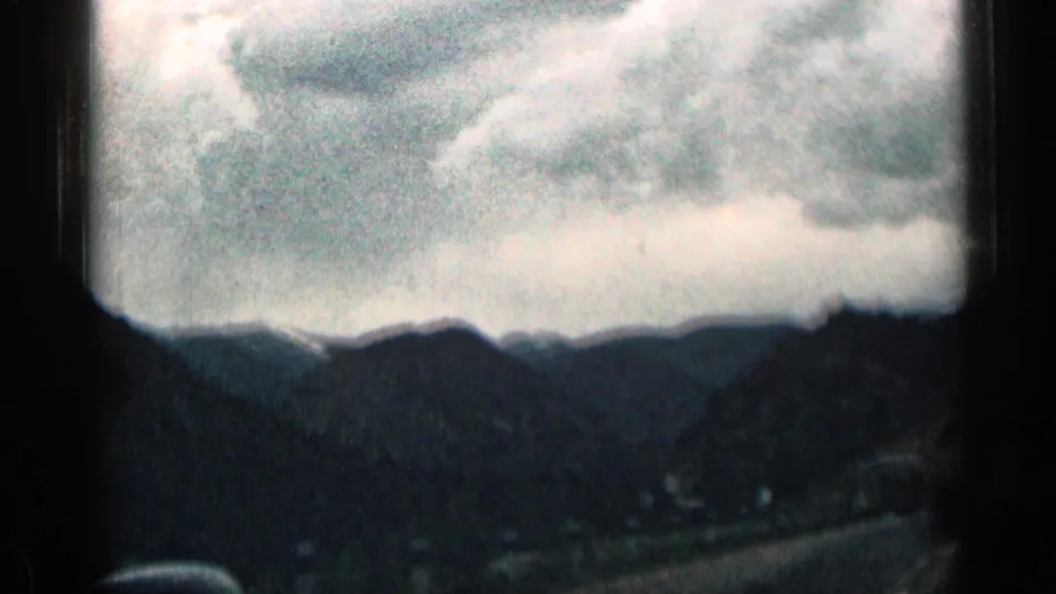 1963 Colorado mountain-road view with cloudy sky, dark ridgelines, and roadside structures in the distance.