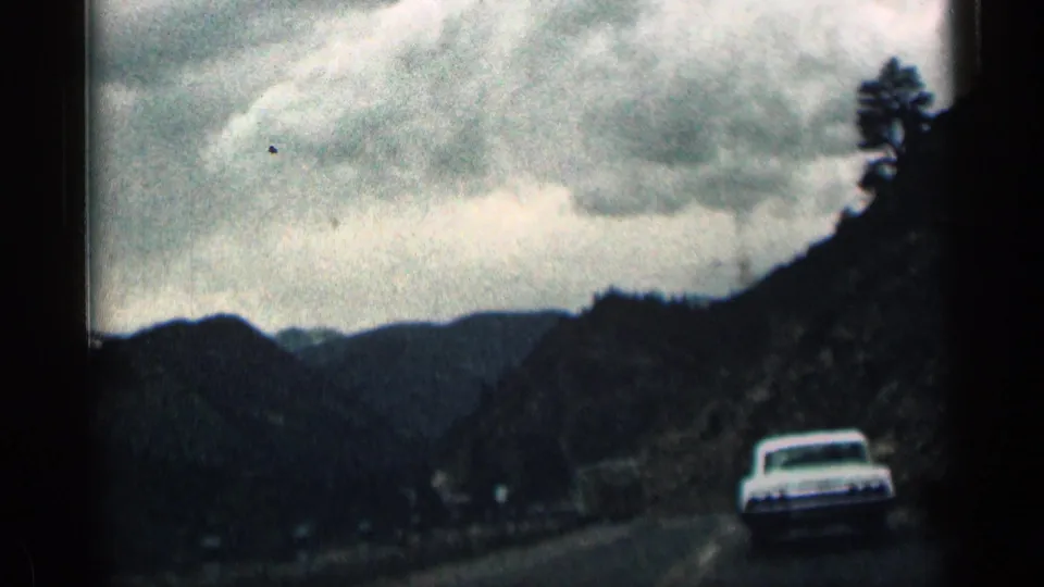 1963 Colorado 8mm frame of a white car ahead on a winding mountain road under an overcast sky.