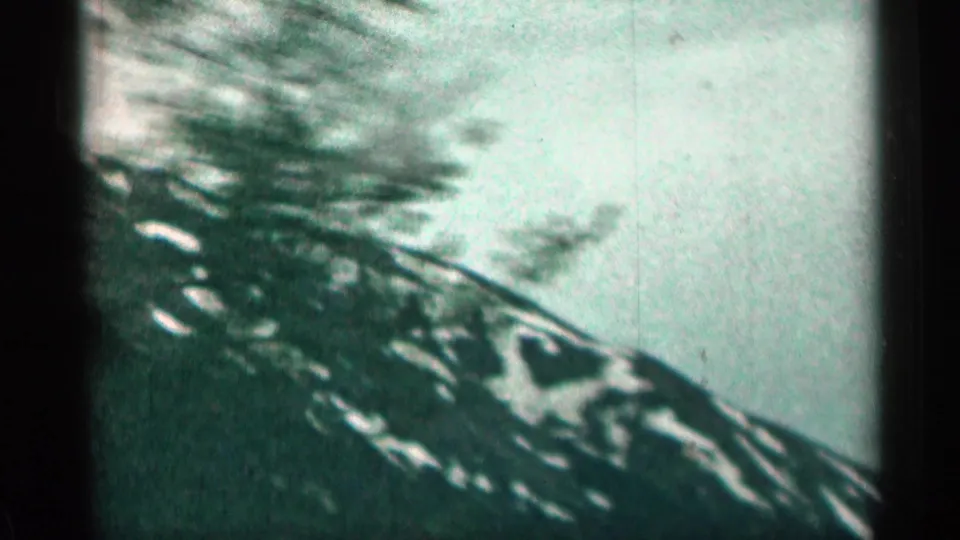 Handheld 1963 Colorado 8mm view with a blurred evergreen in front of a snow-streaked mountain ridge.