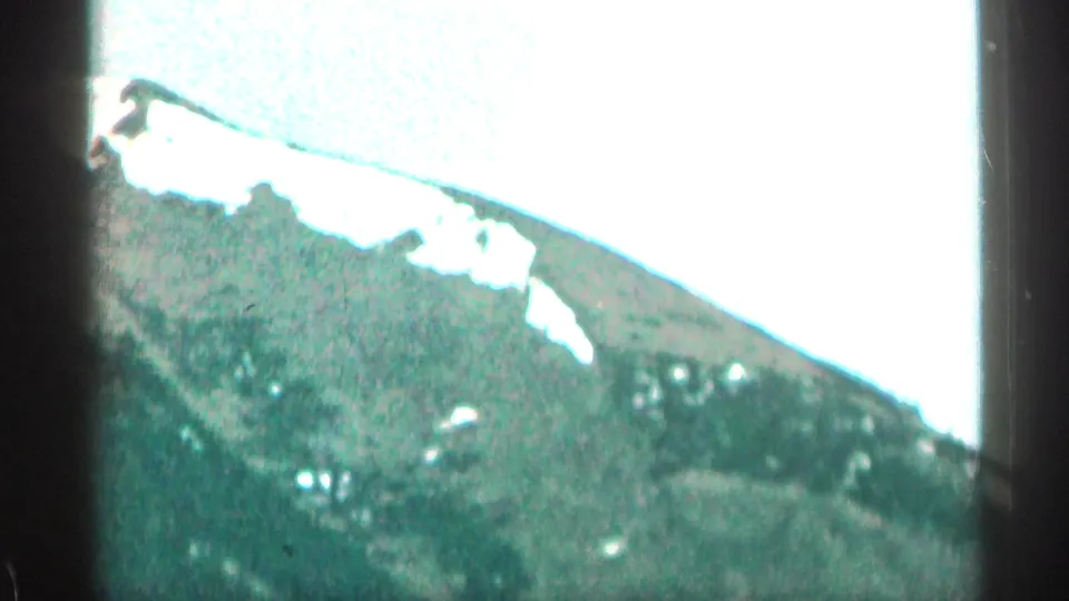 1963 Colorado home-movie frame showing a snow-patched mountain slope and dark forested ridge under an overexposed sky.