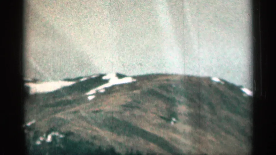 1963 Colorado home-movie frame showing a rounded mountain ridge, scattered snow, and faded color from small-gauge film.