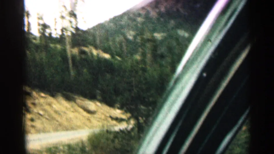 1963 Colorado 8mm frame showing a bend in a mountain road, roadside pines, rocky slope, and the car window frame.