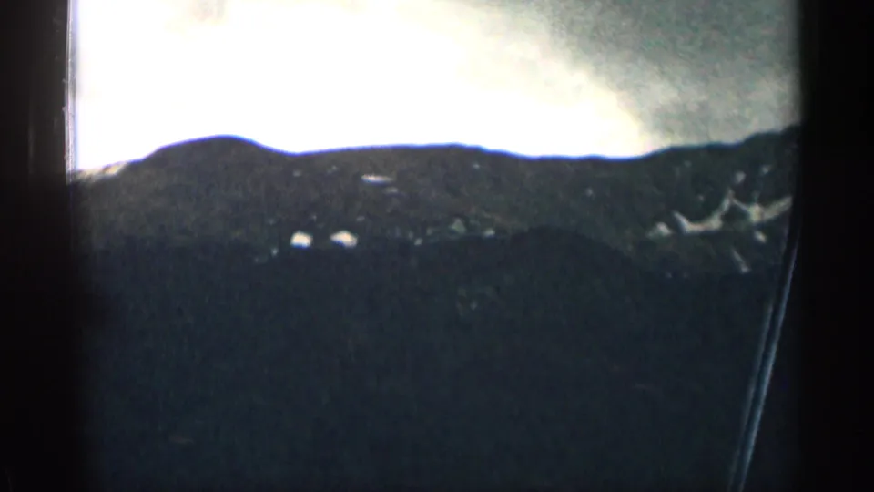 1963 Colorado 8mm frame with a dark mountain ridgeline, overexposed sky, and vehicle window edge during a moving road shot.