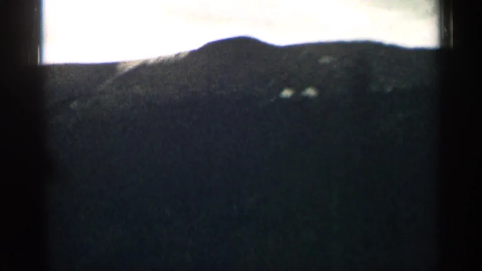 1963 Colorado home-movie frame looking through a car window at a dark mountain slope beneath an overexposed sky.