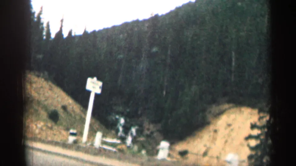 1963 Colorado home-movie view of a mountain road with a roadside sign, evergreen trees, and sloped terrain.