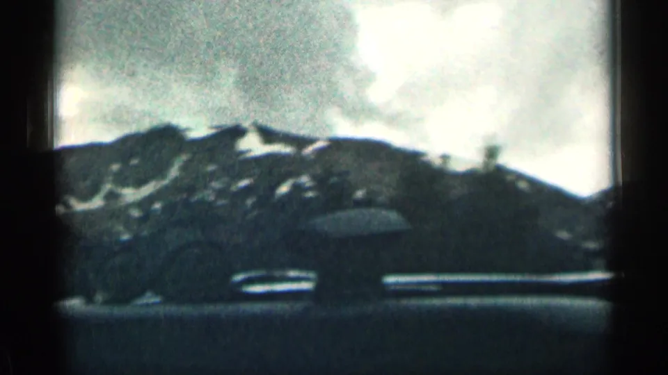 1963 Colorado sightseeing frame with distant snow patches on a mountain ridge seen past the car dashboard.