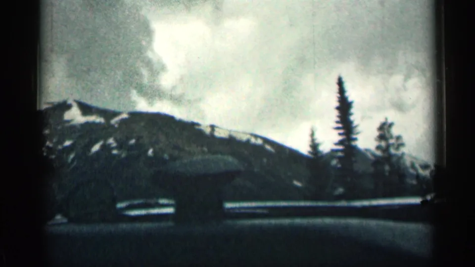1963 Colorado home-movie frame showing snowy mountain slopes and evergreen trees seen through a car window during travel.