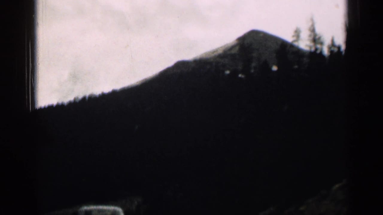 1963 Colorado mountain road footage with a dark ridge and roadside evergreens framed by the car interior.