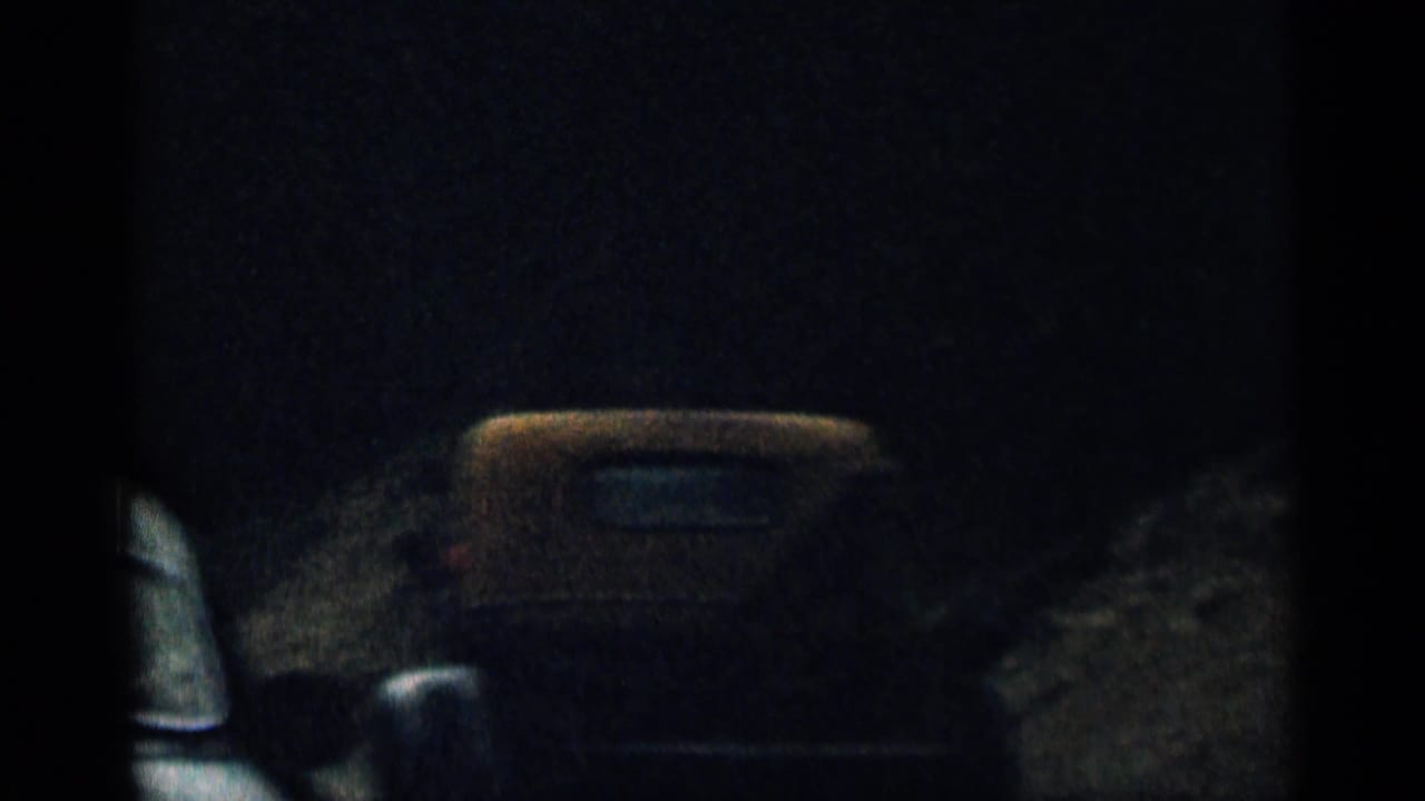 Dark roadside view with a partial car shape at the left edge and shadowed vehicle form ahead.