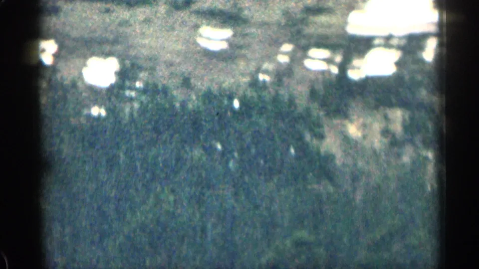 Grainy 1963 Colorado travel frame showing a reflective surface or glare, tree shapes, and overexposed highlights from a moving viewpoint.