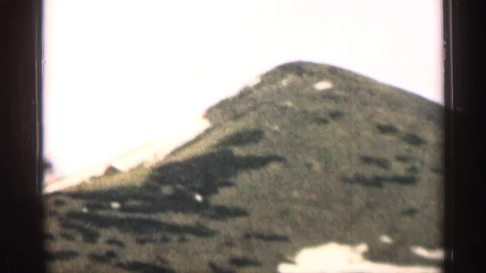 1963 Colorado 8mm frame with a rounded mountain slope, sparse snow or pale patches, and a bright sky near the end of the road-trip clip.