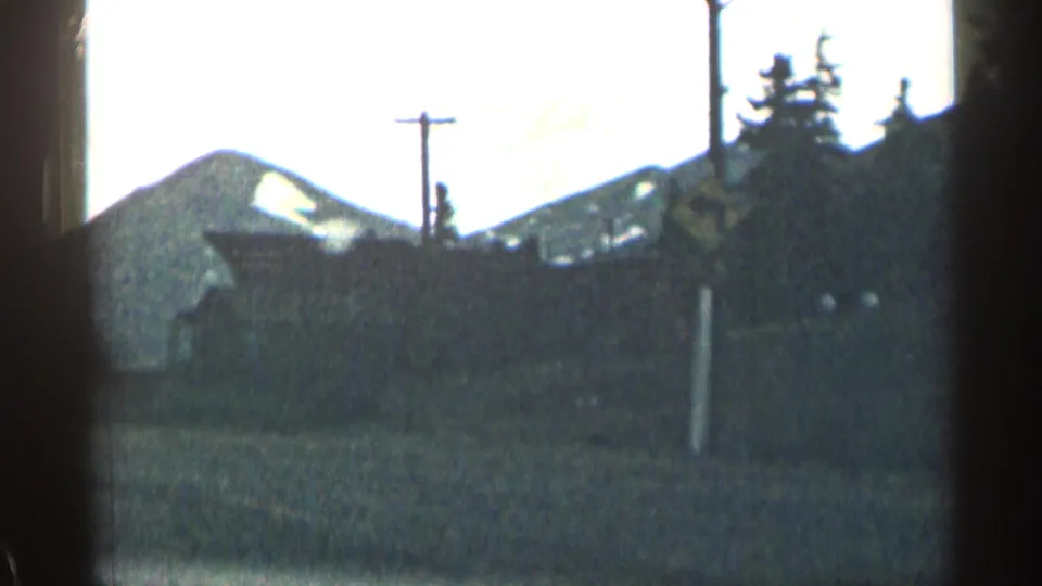 1963 Colorado roadside frame showing a snow-patched mountain slope, evergreen trees, open ground, and a utility pole in soft 8mm focus.