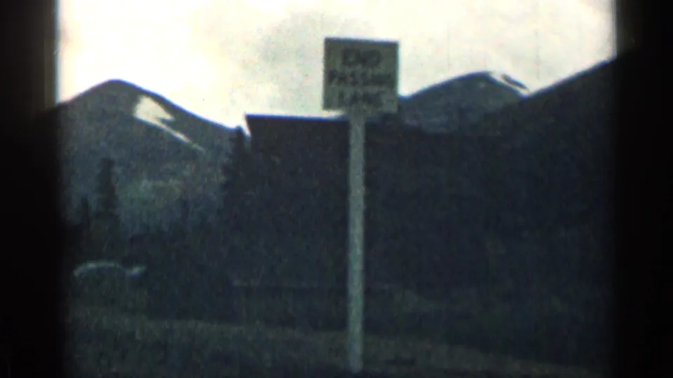 Grainy 1963 Colorado frame with a mountain ridge, snow patches, a roadside signpost, and a dark building or post in the foreground.