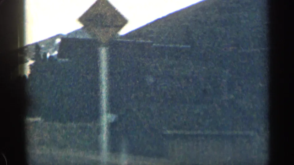 A large sloped-roof building and fence or post appear below a Colorado mountain ridge in soft, grainy 8mm footage.