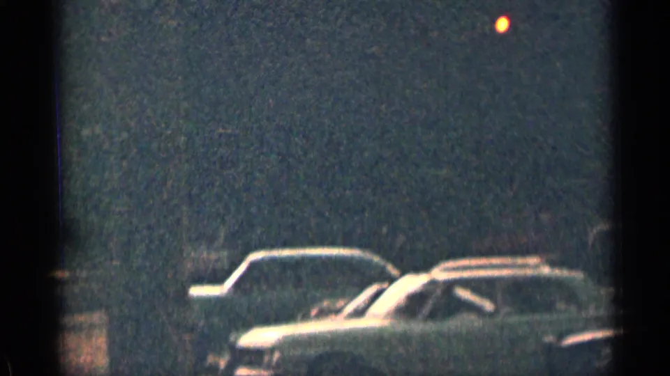 Grainy 1963 Colorado 8mm frame showing a dim dirt parking area with partial classic American cars near the lower edge.