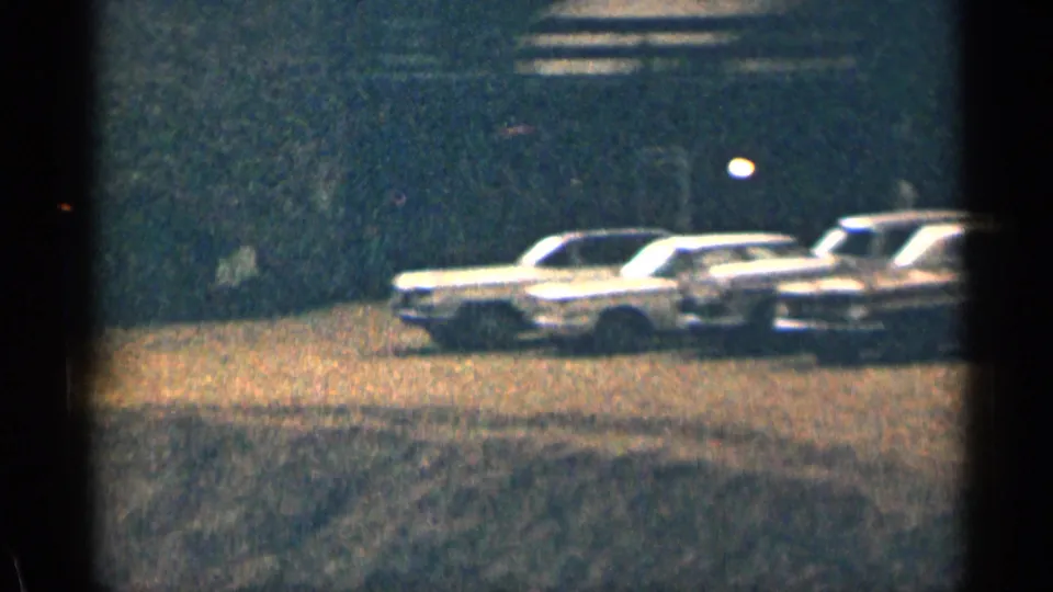 Low-light 1963 Colorado frame showing several classic cars parked on a dirt lot beneath dim lights and strong film grain.
