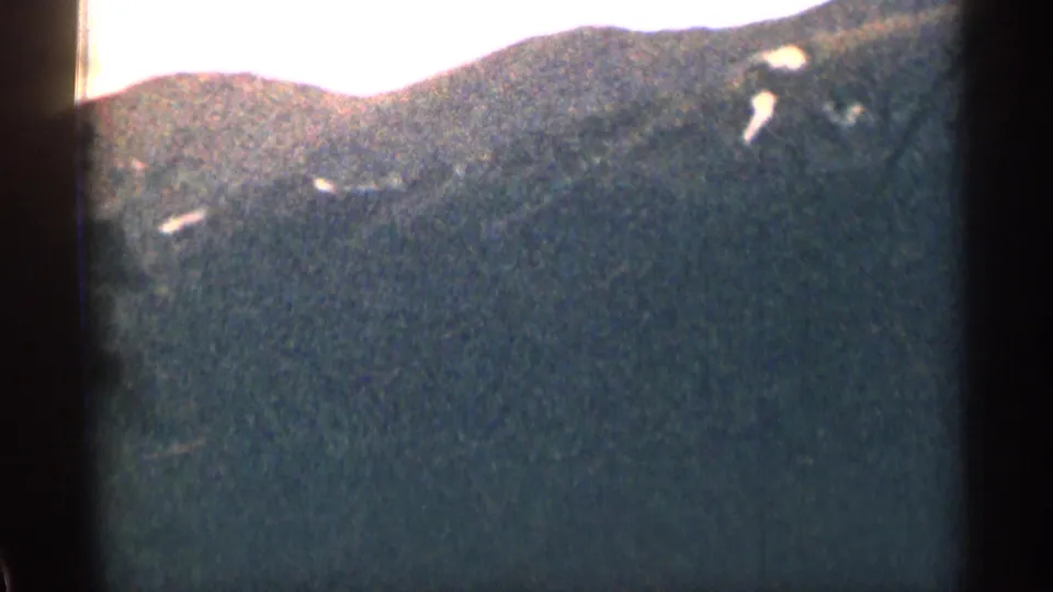 Faded 8mm frame from Colorado showing a dark mountain or wooded ridge under bright sky with visible film wear and grain.