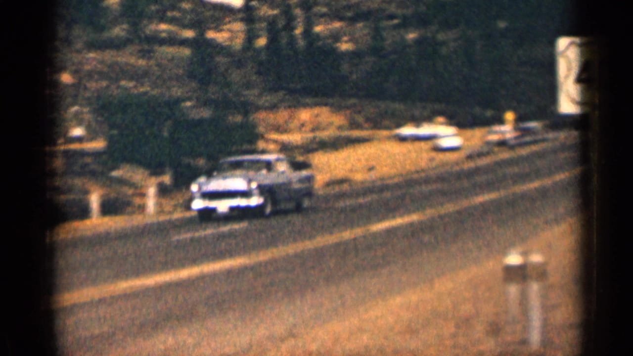 Light-colored vintage sedan driving along a rural two-lane highway.
