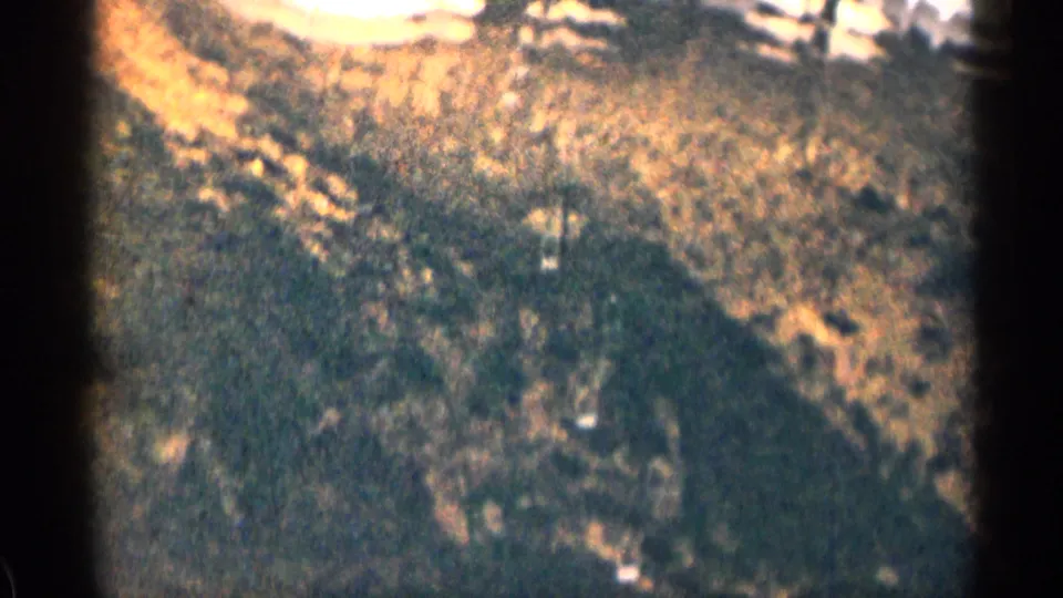 1963 Colorado 8mm frame showing a sunlit rocky mountain slope, dark trees, and bright patches that may be melting snow.