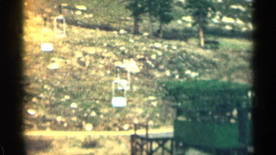 1963 Colorado ski-area view with lift chairs, support towers, rocks, and trees on a hillside.