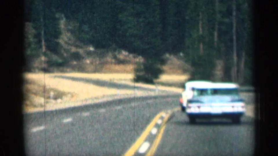 1963 Colorado road view with a light-colored period car ahead on a curving two-lane route through evergreens.