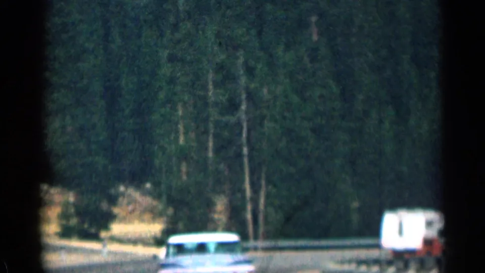 1963 Colorado 8mm frame showing a road through dense pine forest with a car and small light-colored vehicle partly visible.