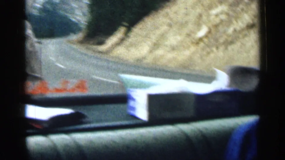 Interior car-window view of a vehicle ahead on a mountain road in Colorado, with road edge and hillside visible.