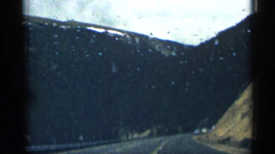 1963 Colorado road-trip frame with a mountain road, steep slope, snow-dusted ridge, and cloudy sky.