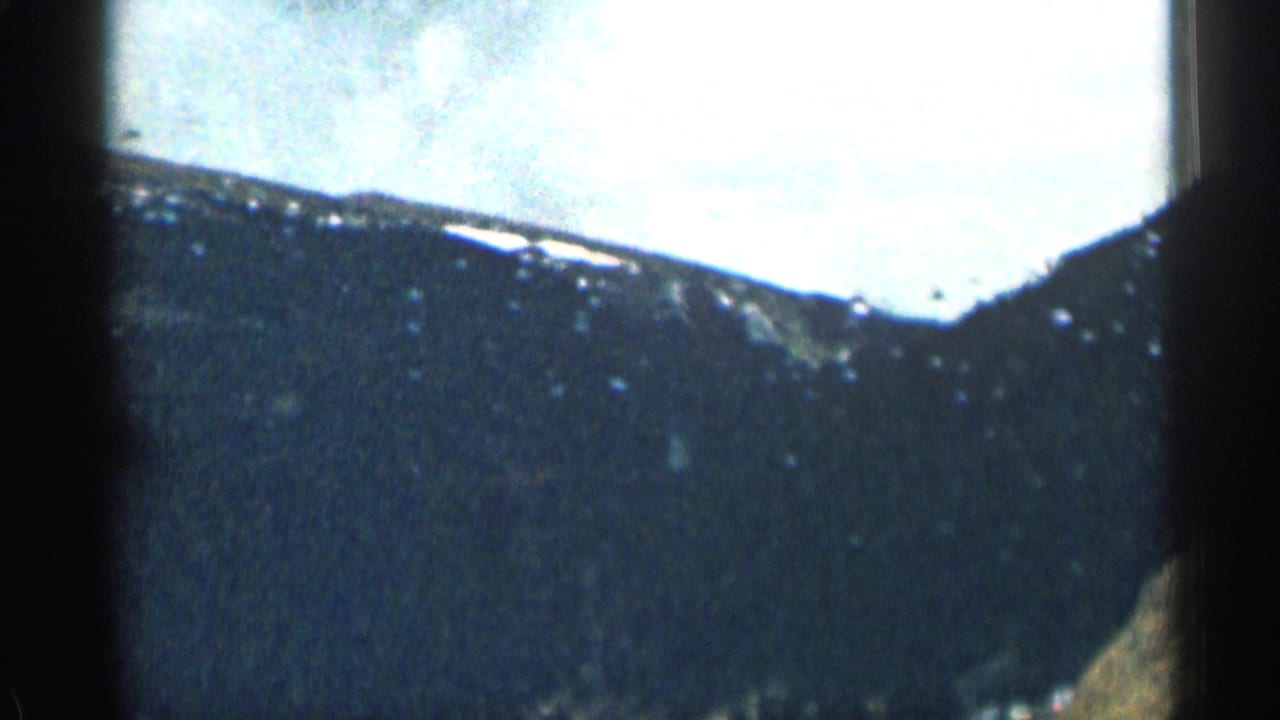 1963 Colorado 8mm frame from a moving-car perspective, with dark mountain terrain, bright sky, soft focus, and heavy film grain visible through the windshield.