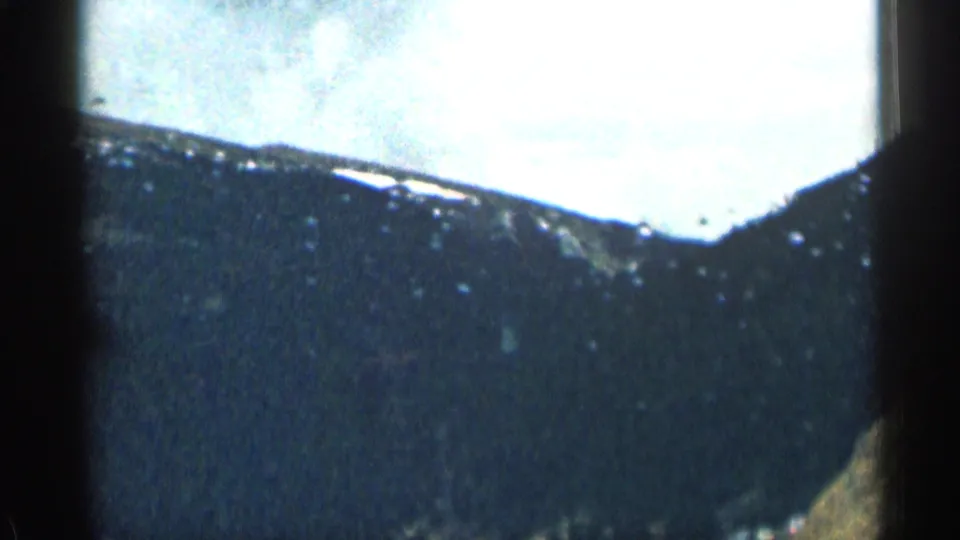 1963 Colorado 8mm frame from a moving-car perspective, with dark mountain terrain, bright sky, soft focus, and heavy film grain visible through the windshield.