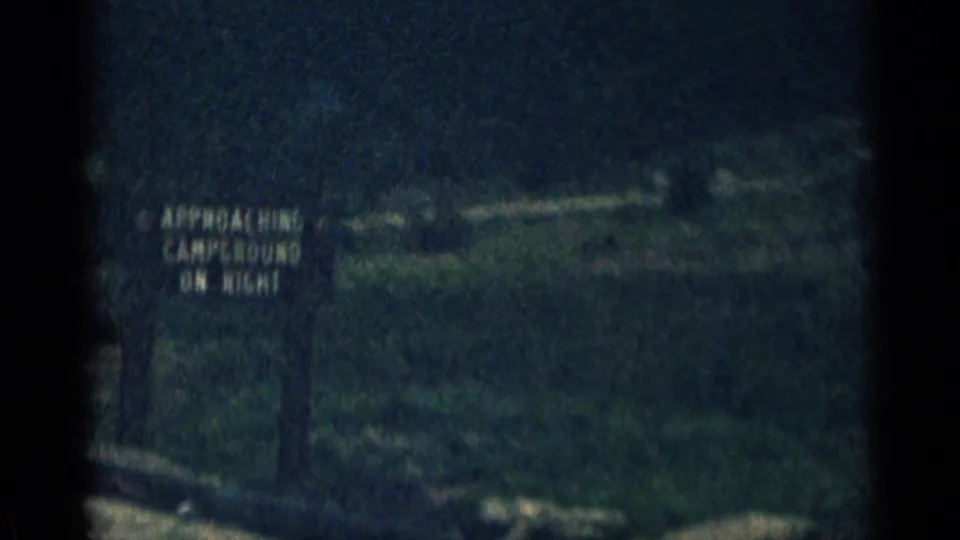 1963 Colorado roadside frame with a partially legible campground approach sign beside rugged mountain terrain, seen in soft, grainy 8mm color.
