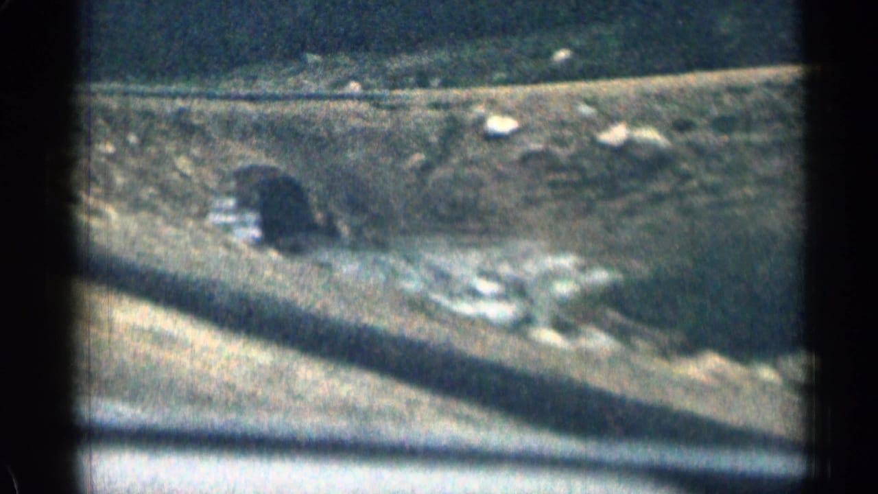 1963 Colorado 8mm view of a small bridge or culvert over moving water, with a road edge and rocky roadside landscape in the foreground.